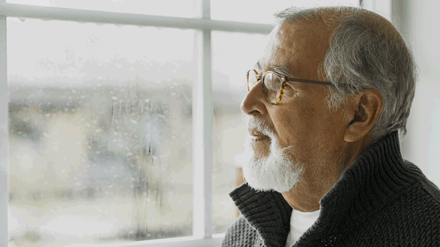 A sad elderly gentleman looking out through a window. Photo. 