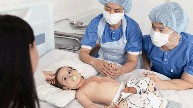A photo of a toddler with severe burns in a hospital bed. A mother and two doctors are tending to the child. 