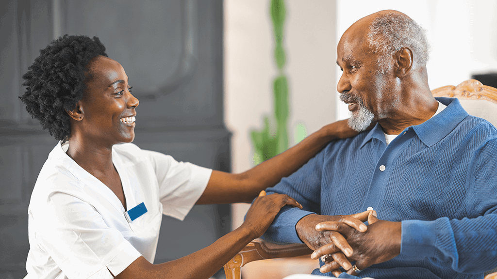A photo of a patient sitting in a chair smiling at a nurse, the nurse is crouched down on eye level with the patient and smiling back at them. 
