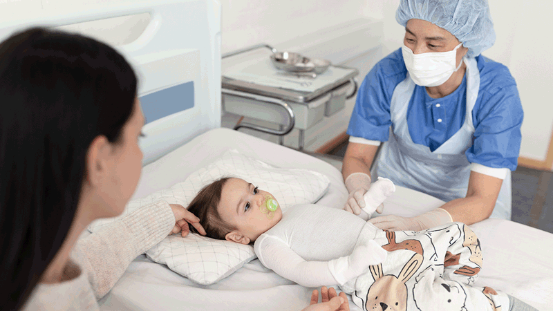 A photo of a toddler wearing burn dressings on all of its upper body. The toddler is tended to by a mother and a doctor. 